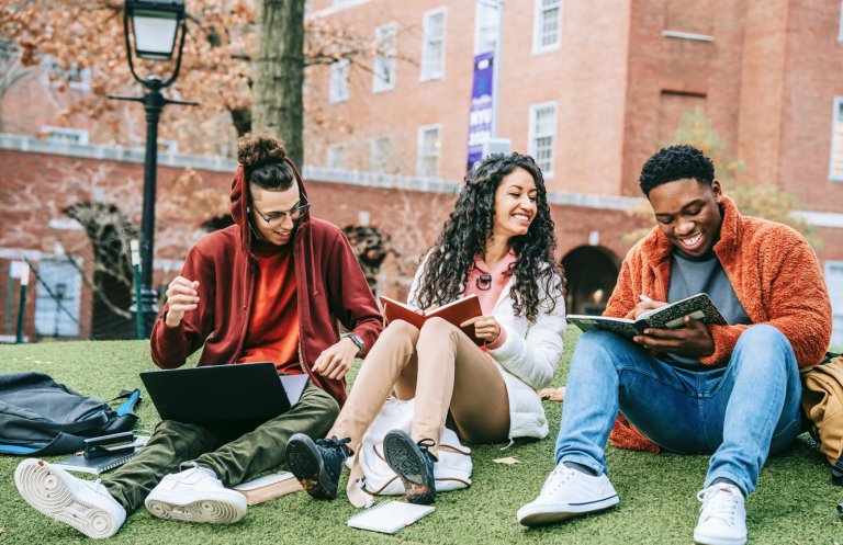 Three college-aged students study together on a campus lawn