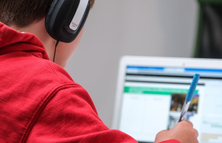 A student takes notes and listens to audio while working on their laptop.